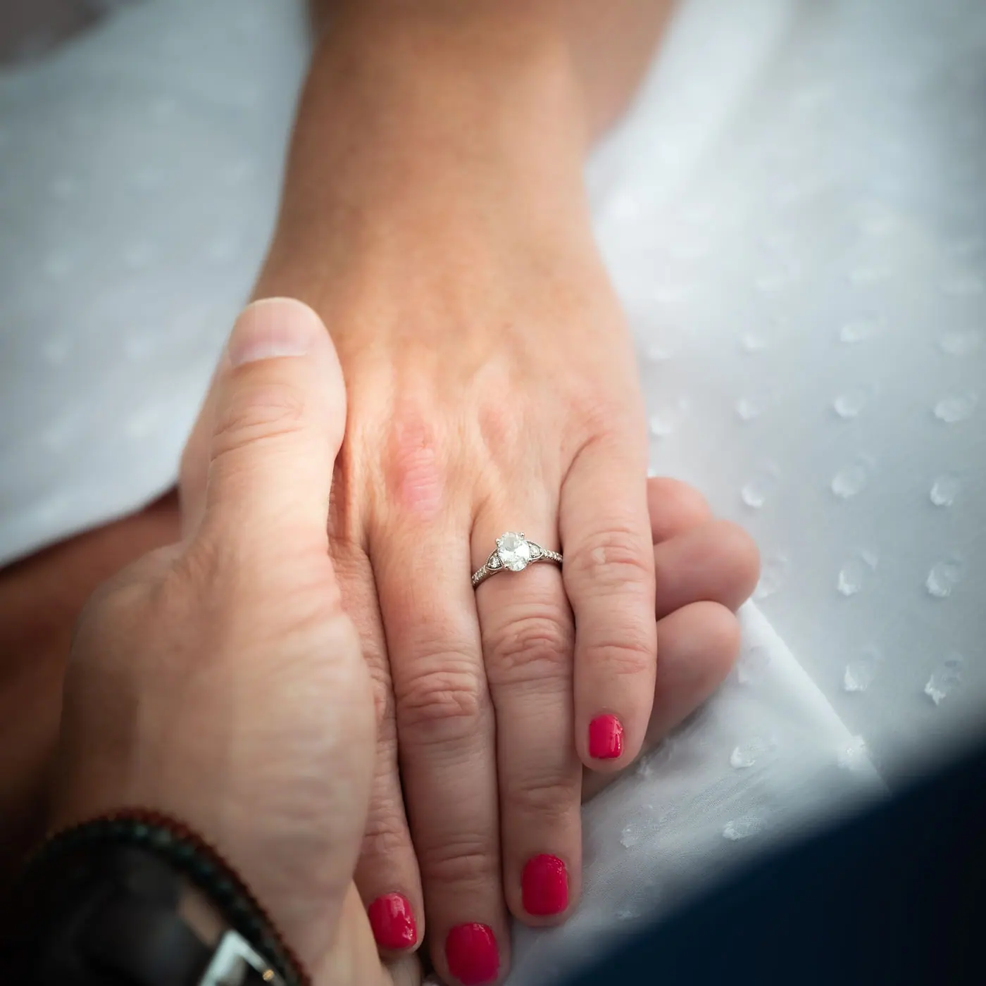 Hands with red nails, one wearing a diamond solitaire engagement ring