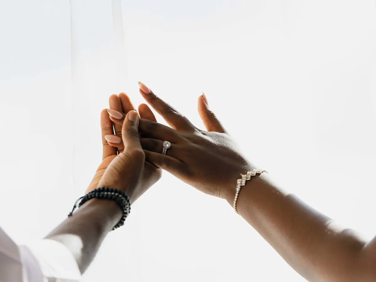 Two hands touching in window light, one wearing a diamond ring and a diamond line bracelet
