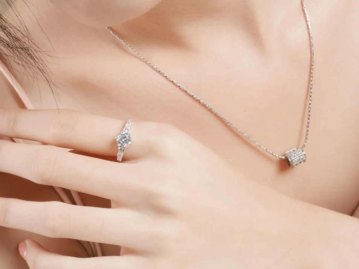 Hand at the collarbone wearing a round diamond solitaire ring and a fine diamond necklace