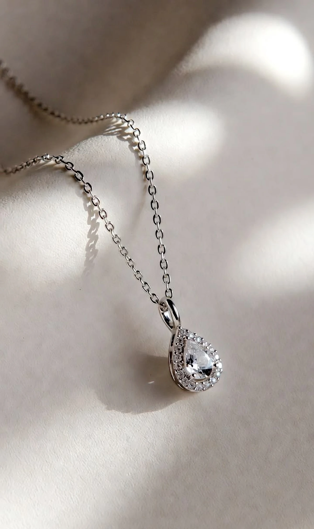 Pear-shaped diamond halo pendant on a fine white-gold chain resting on soft fabric